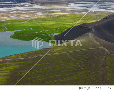 A vibrant turquoise lake surrounded by green vegetation, dark rugged hills with trails, and braided rivers flowing across a flat plain in Iceland. 133108025