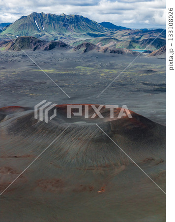 A reddish brown volcanic crater in Iceland surrounded by barren terrain, with rugged mountains, green vegetation, and snow under a partly cloudy sky. A reddish brown volcanic crater in Iceland surrounded by barren terrain, with rugged mountains, green vegetation, and snow under a partly cloudy sky. 133108026