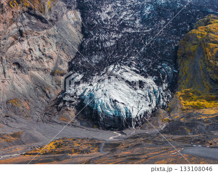 A glacier in Iceland with icy blue hues contrasts with dark volcanic rock and yellow moss. A winding path leads to the glacier, showcasing its scale and terrain. 133108046