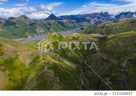 Aerial view of a green valley in Iceland with deep ravines, rolling hills, and a braided river. Snow capped peaks and glaciers are visible under a blue sky. 133108047