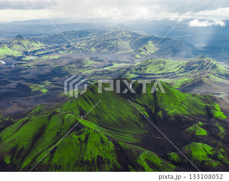 Aerial view of Iceland's volcanic terrain with a green mountain, dark soil, rolling hills, and valleys under soft light and scattered clouds. Aerial view of Iceland's volcanic terrain with a green mountain, dark soil, rolling hills, and valleys under soft light and scattered clouds. 133108052