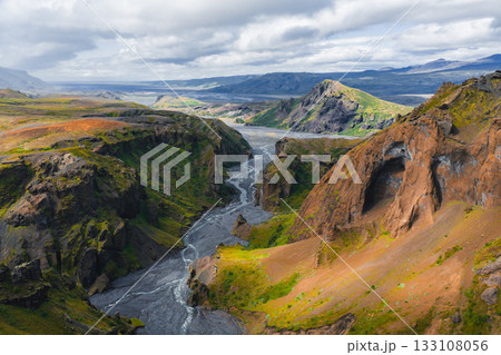 Mulagljufur Canyon in Iceland features dramatic cliffs, green vegetation, a winding river, and a weathered rock formation under a partly cloudy sky. 133108056