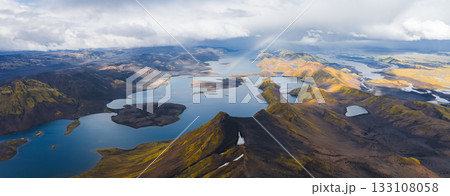 Aerial view of interconnected lakes in Iceland's highlands, surrounded by moss covered mountains, volcanic terrain, and a faint rainbow in the sky. 133108058