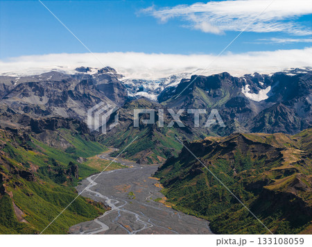 A winding river in Iceland flows through a lush green valley surrounded by cliffs and jagged mountains, with snow capped peaks and glaciers in the background. 133108059