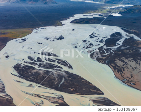 Aerial view of a pale blue glacial river weaving through dark volcanic sands, with distant hills and mountains in Iceland's rugged landscape. Aerial view of a pale blue glacial river weaving through dark volcanic sands, with distant hills and mountains in Iceland's rugged landscape. 133108067
