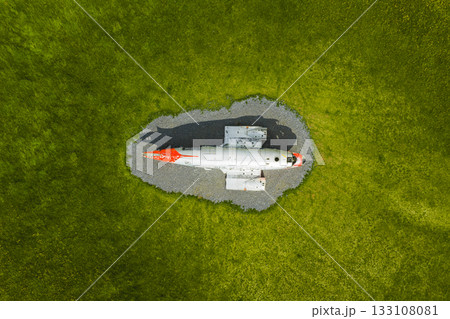 An aerial view of a weathered red and white Abandoned U.S. Navy Douglas C 117D aircraft on gravel, surrounded by vibrant green grass in Iceland's dramatic natural landscape. 133108081