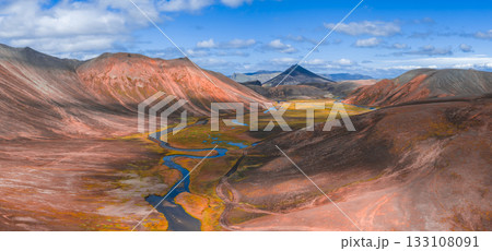 Aerial view of colorful rhyolite mountains, a winding river, and lush green valley under a bright blue sky with a triangular peak in the background. 133108091