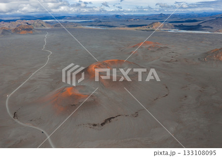 Aerial view of a barren volcanic terrain in Iceland, featuring reddish orange craters, a winding dirt road, distant mountains, and scattered clouds. Aerial view of a barren volcanic terrain in Iceland, featuring reddish orange craters, a winding dirt road, distant mountains, and scattered clouds. 133108095