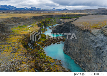 Sigoldugljufur Canyon features moss covered cliffs, cascading waterfalls, a turquoise river, rocky terrain, and distant snow capped mountains under cloudy skies. 133108135