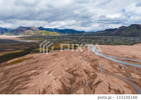 Aerial view of a braided river system in Iceland on a reddish brown plain, surrounded by rugged, multicolored mountains with green vegetation under a cloudy sky. 133108166