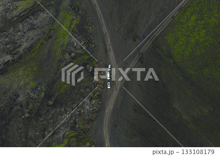 Aerial image of Iceland's volcanic landscape with green moss patches, featuring three vehicles at a dirt road intersection in a vast, isolated area. 133108179