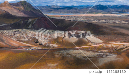 Aerial view of a volcanic crater in Iceland, surrounded by red, orange, and yellow hills. A dirt road with parked vehicles cuts through rugged terrain. Aerial view of a volcanic crater in Iceland, surrounded by red, orange, and yellow hills. A dirt road with parked vehicles cuts through rugged terrain. 133108180