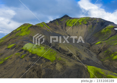 A steep volcanic mountain in Iceland features dark rocky slopes covered with vibrant green moss under a partly cloudy sky, creating a dramatic scene. 133108189