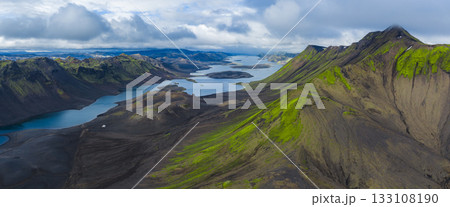 Aerial view of elongated lakes amid volcanic terrain and green moss covered mountains in Iceland, framed by dramatic cloud cover and sharp ridges. 133108190