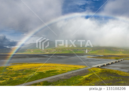 A vibrant rainbow arches over a river spanned by a long bridge. A waterfall cascades down a green hillside in the background of the lush Icelandic landscape. 133108206