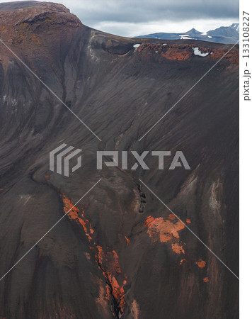 Dark ash covered slopes with orange and red streaks, steep terrain with crevices, and snow capped peaks under a cloudy sky in Iceland. 133108227