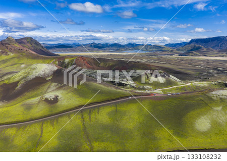 Aerial view of a volcanic crater surrounded by green moss covered hills, dark volcanic soil, a winding road, distant mountains, and a serene lake. Aerial view of a volcanic crater surrounded by green moss covered hills, dark volcanic soil, a winding road, distant mountains, and a serene lake. 133108232