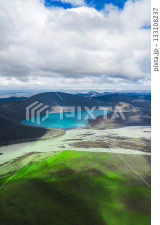 A vibrant turquoise lake surrounded by dark volcanic hills and moss covered terrain, with a partly cloudy sky adding depth to the Icelandic landscape. 133108237