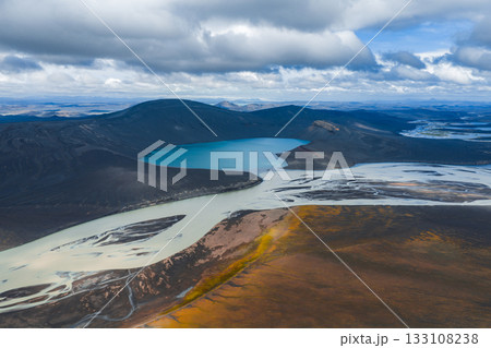 A vivid blue lake surrounded by dark volcanic hills, with a braided river system flowing through rugged terrain. Earthy tones and yellow vegetation are visible. 133108238