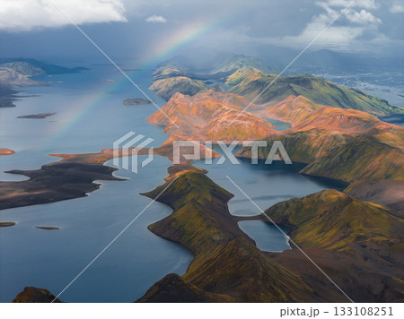 Aerial view of Iceland's rhyolite mountains with vivid colors, interconnected lakes, and a rainbow under a partly cloudy sky in the Landmannalaugar region. Aerial view of Iceland's rhyolite mountains with vivid colors, interconnected lakes, and a rainbow under a partly cloudy sky in the Landmannalaugar region. 133108251