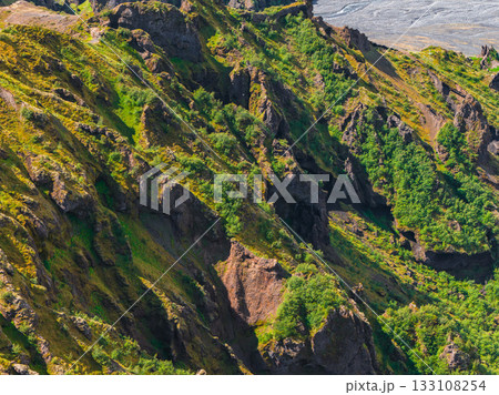 Lush green cliffs with steep rocky formations and deep crevices, set against a flat gray glacial outwash plain in Iceland's dramatic landscape. Lush green cliffs with steep rocky formations and deep crevices, set against a flat gray glacial outwash plain in Iceland's dramatic landscape. 133108254