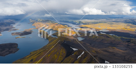 Aerial view of Iceland's highlands with vibrant hills, deep blue lakes, volcanic rock, green moss, snow patches, and a faint rainbow under cloudy skies. Aerial view of Iceland's highlands with vibrant hills, deep blue lakes, volcanic rock, green moss, snow patches, and a faint rainbow under cloudy skies. 133108255