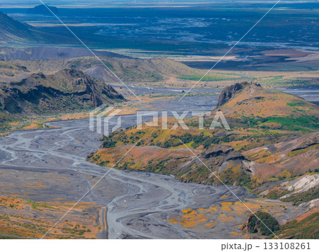 A braided river winds through a valley with volcanic soil, green vegetation, and orange moss, surrounded by hills and rocky formations in Iceland. A braided river winds through a valley with volcanic soil, green vegetation, and orange moss, surrounded by hills and rocky formations in Iceland. 133108261
