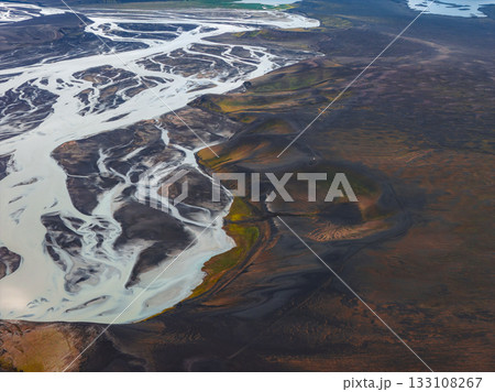 Braided river channels flow through dark volcanic terrain in Iceland, with small craters and green vegetation adding texture to the dramatic scene. 133108267