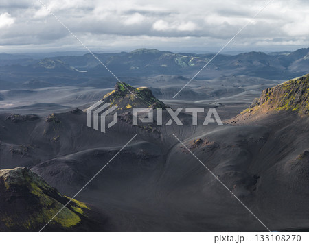 Dark volcanic terrain with vibrant green moss, rugged hills, and distant mountains under a cloudy sky, creating a dramatic Icelandic scene. 133108270