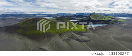 Aerial view of moss covered volcanic hills, craters, and a blue crater lake in Iceland. Snow capped mountains and distant lakes appear under a cloudy sky. Aerial view of moss covered volcanic hills, craters, and a blue crater lake in Iceland. Snow capped mountains and distant lakes appear under a cloudy sky. 133108277