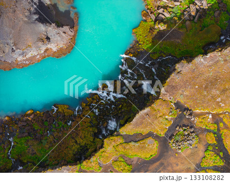 Turquoise river winds through rugged volcanic terrain in Iceland, cascading over dark rocks with small waterfalls, surrounded by moss and earthy tones. 133108282