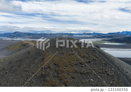 Visitors stand on a rocky volcanic ridge in Iceland, surrounded by plains, winding rivers, distant mountains, and patches of green vegetation under an overcast sky. Visitors stand on a rocky volcanic ridge in Iceland, surrounded by plains, winding rivers, distant mountains, and patches of green vegetation under an overcast sky. 133108350