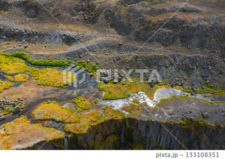 An aerial view of a rugged Icelandic landscape with a narrow waterfall cascading over a cliff, bordered by basalt columns, moss, and golden vegetation. 133108351