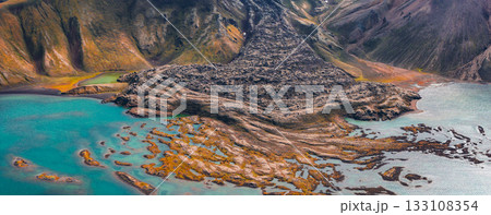 Aerial view of a volcanic terrain in Iceland with a lava flow leading into a turquoise lake, surrounded by multicolored hills and green moss. 133108354