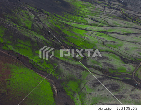 Aerial view of Iceland's volcanic terrain with green moss, dark soil, undulating hills, valleys, and a small road with a white vehicle visible. Aerial view of Iceland's volcanic terrain with green moss, dark soil, undulating hills, valleys, and a small road with a white vehicle visible. 133108355