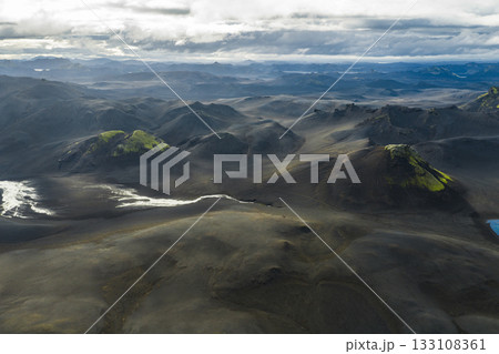 Dramatic aerial view of Iceland's volcanic highlands with dark soil, green moss covered hills, a winding river, and a moody cloudy sky. 133108361