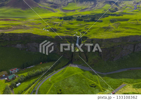Aerial view of a waterfall in Iceland flowing from a cliff into a lush valley, surrounded by greenery, winding paths, and small buildings below. Aerial view of a waterfall in Iceland flowing from a cliff into a lush valley, surrounded by greenery, winding paths, and small buildings below. 133108362