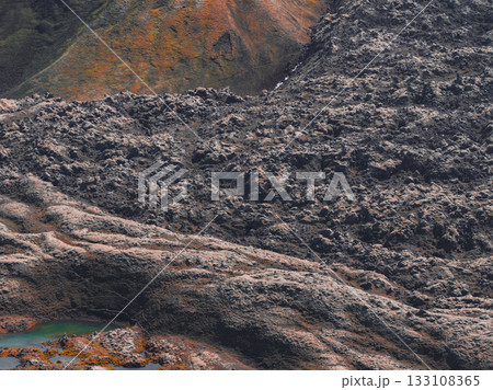 Rugged volcanic terrain in Iceland with dark lava fields, earthy orange and green slopes, and a vivid green and orange pool in the foreground. 133108365