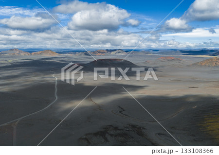 A barren volcanic terrain in Iceland with ash covered ground, red craters, a winding dirt road, and a cloudy sky casting dynamic shadows. 133108366