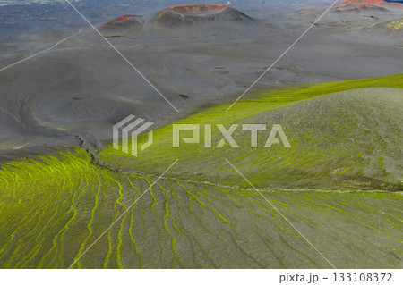 Aerial view of a volcanic terrain in Iceland with dark slopes, green moss, red tinted craters, and a faint road cutting through the remote area. 133108372