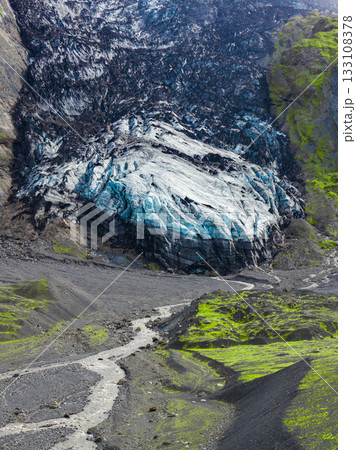 A glacier in Iceland features icy blue and white streaked with volcanic ash, surrounded by mossy slopes and a winding stream in a rugged landscape. A glacier in Iceland features icy blue and white streaked with volcanic ash, surrounded by mossy slopes and a winding stream in a rugged landscape. 133108378