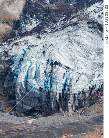 A glacier in Iceland features vivid blue ice streaks and dark crevices, surrounded by rocky terrain with orange vegetation and intricate ice patterns. A glacier in Iceland features vivid blue ice streaks and dark crevices, surrounded by rocky terrain with orange vegetation and intricate ice patterns. 133108382