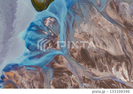 Braided river system with vibrant blue glacial waters flowing through earthy tones, featuring a small green island and intricate geological patterns. 133108398