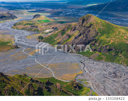 Aerial view of a rocky Icelandic valley with a winding river, dark volcanic soil, green vegetation, jagged hills, and distant mountains. Aerial view of a rocky Icelandic valley with a winding river, dark volcanic soil, green vegetation, jagged hills, and distant mountains. 133108422
