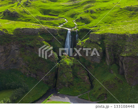 Aerial view of a waterfall in Iceland surrounded by green moss and grass, flowing into a gorge with visible walking paths and a parking area below. 133108448