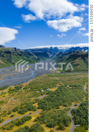Aerial view of a green valley in Iceland with a winding river, rugged mountains, glaciers, scattered tents, vehicles, and a bright blue sky. Aerial view of a green valley in Iceland with a winding river, rugged mountains, glaciers, scattered tents, vehicles, and a bright blue sky. 133108450