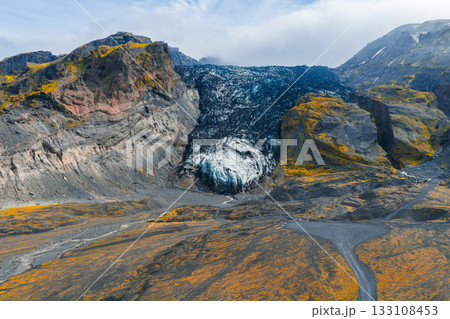 A glacier in Iceland with icy blue tones lies between volcanic cliffs covered in golden moss. Tiny figures and pathways highlight the scale of the dramatic landscape. 133108453