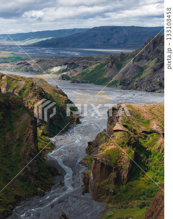 Steep cliffs with green vegetation surround a glacial river in Mulagljufur Canyon, Iceland. Volcanic soil and distant mountains complete the scene. 133108458