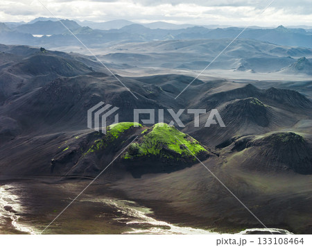 Aerial view of Iceland's volcanic terrain featuring dark soil, a vibrant green moss covered hill, distant mountain ranges, and a cloudy sky. 133108464