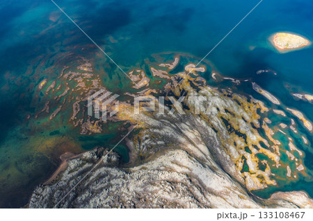 Aerial view of deep blue waters with golden and green patterns of algae or minerals, featuring a small island in the upper right corner in Iceland. Aerial view of deep blue waters with golden and green patterns of algae or minerals, featuring a small island in the upper right corner in Iceland. 133108467